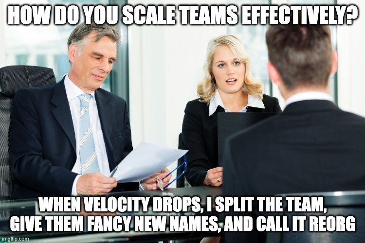 The image shows a job interview setting with three people in a modern office. A middle-aged man in a suit is holding a folder and smiling, while a young woman in a blazer looks serious. They are interviewing a man whose back is facing the viewer. The meme text reads: 

**Top text:** "HOW DO YOU SCALE TEAMS EFFECTIVELY?"  
**Bottom text:** "WHEN VELOCITY DROPS, I SPLIT THE TEAM, GIVE THEM FANCY NEW NAMES, AND CALL IT REORG."

The meme satirizes how some companies handle team management by reorganizing teams rather than solving productivity challenges.